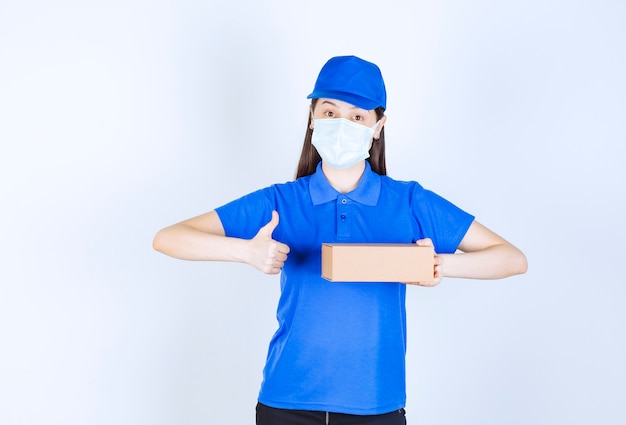 Young deliverywoman in medical mask holding carton box and giving thumbs up. 