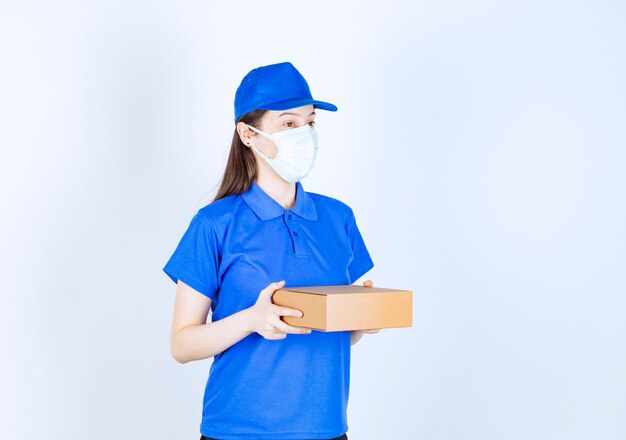 Young deliverywoman in medical mask holding carton box on white background. 