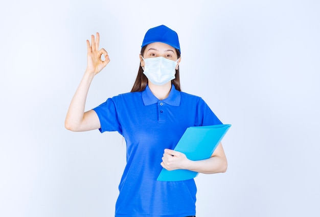 Young deliverywoman in medical mask holding clipboard and giving ok sign. 