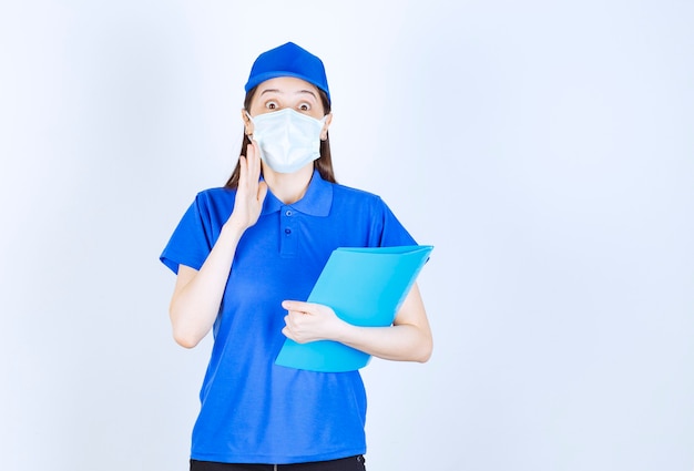 Young deliverywoman in medical mask holding clipboard on white background. 