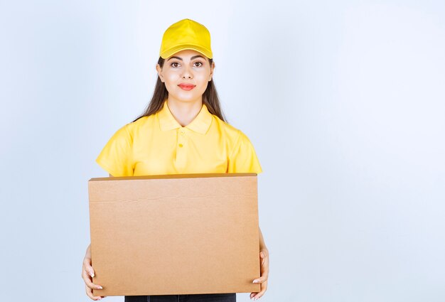 Young deliverywoman in yellow cap holding carton package over white wall.