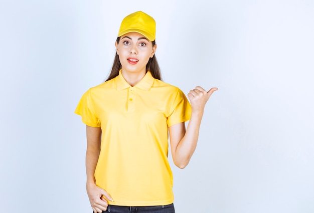 Young deliverywoman in yellow t-shirt and cap pointing at somewhere on white.