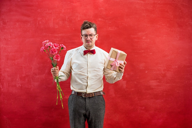 Молодой разочарованный забавный человек с цветами и подарком на красной студии
