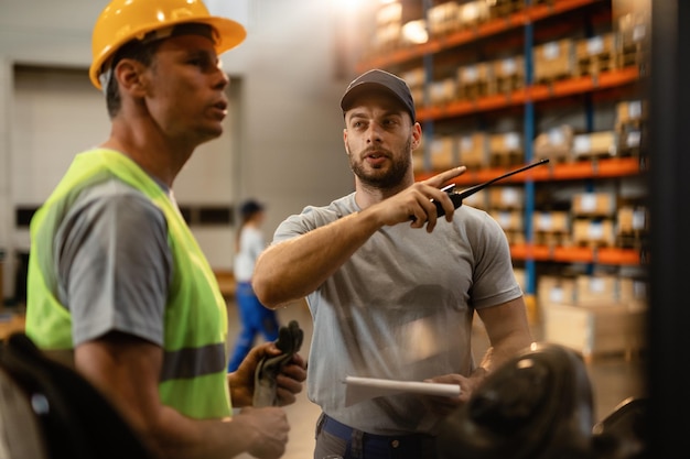 Young dispatcher communicating about delivery schedule with warehouse worker in factory warehouse