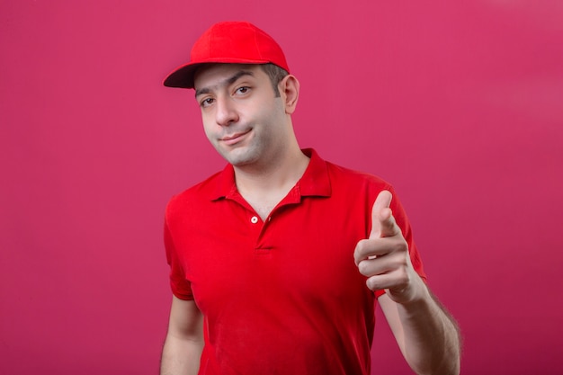 빨간색 폴로 셔츠와 격리 된 분홍색 배경 위에 카메라에 손가락을 가리키는 모자에 젊은 불쾌 배달 남자