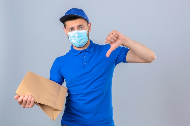 Молодой недовольный курьер в синей рубашке поло и кепке в защитной медицинской маске, стоящий с бумажным пакетом, показывая большой палец вниз на изолированном белом фоне