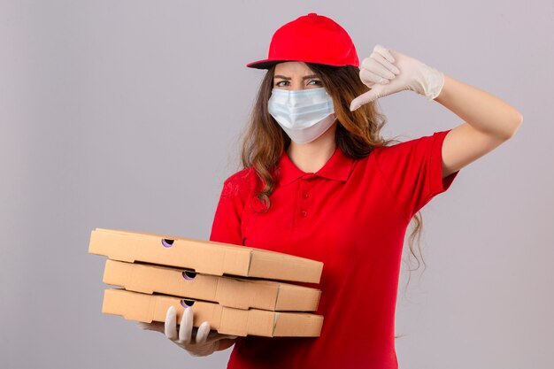 Молодая недовольная женщина-доставщик с вьющимися волосами в красной рубашке поло и кепке в медицинской защитной маске и перчатках стоит с коробками для пиццы, демонстрируя неприязнь на изолированном белом фоне