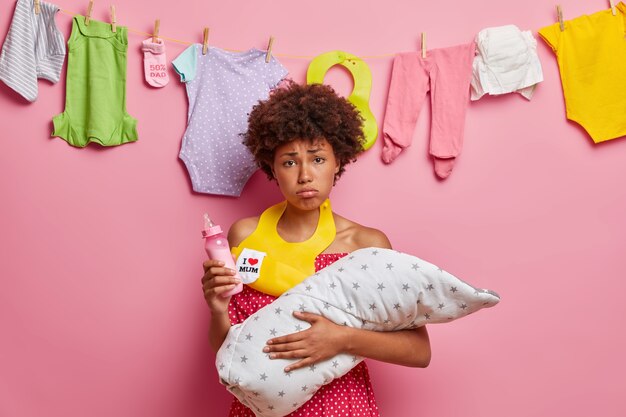 Young displeased tired mother holds baby wrapped in blanket holds feeding bottle wears bib around neck going to feed newborn has much work about house. Mom nursing small daughter. One parent family