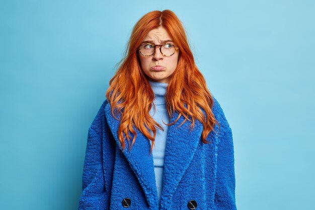 Young displeased woman purses lips and looks with sullen expression aside dressed in turtleneck and blue winter coat.