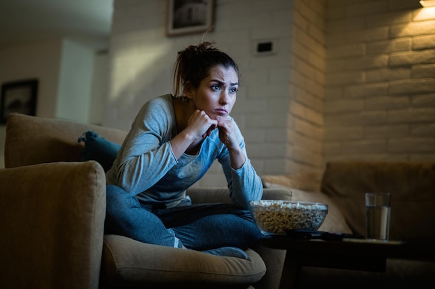 Giovane donna dispiaciuta seduta in poltrona e guardando la tv a tarda notte a casa
