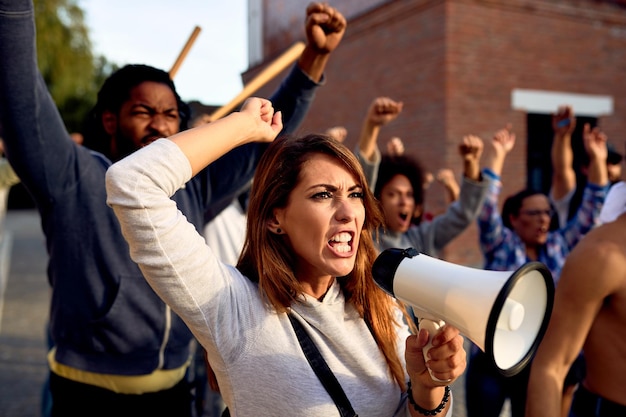 Young displeased woman using megaphone and shouting while protesting with group of people on city streets