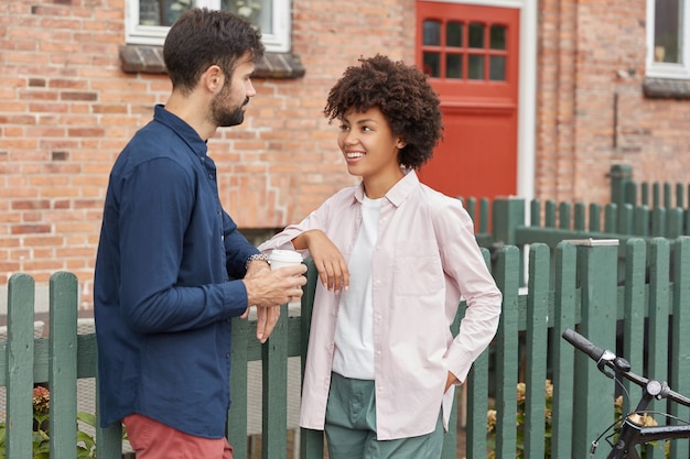 Young diverse couple meet in rural street, stand near green fence and brick house, have positive talk