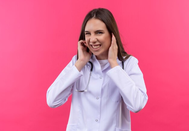 孤立したピンクの壁に聴診器の医療用ガウンの閉じた耳を身に着けている若い医者の女の子