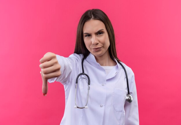 Молодая девушка-врач в медицинском халате со стетоскопом, опустив большой палец на изолированную розовую стену