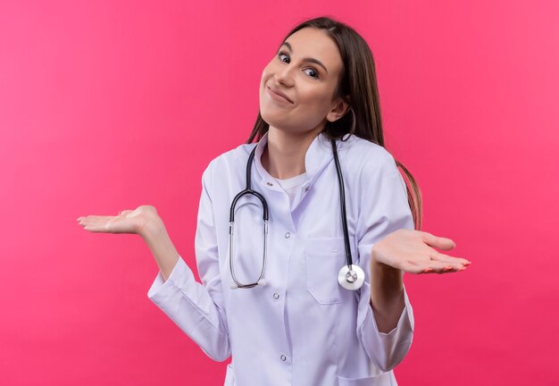  young doctor girl wearing stethoscope medical gown spreads hands on isolated pink wall