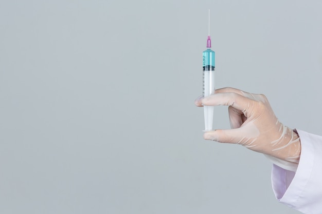 Young doctor is  holding  hypodermic syringe with rubber gloves on gray wall.