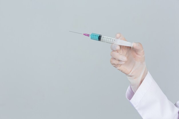 Young doctor is  holding  hypodermic syringe with rubber gloves on gray wall.