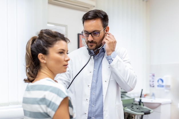 若い医者は聴診器を使用して患者の心拍を聞きます女性の患者に健康診断を与える男性の医者のショット