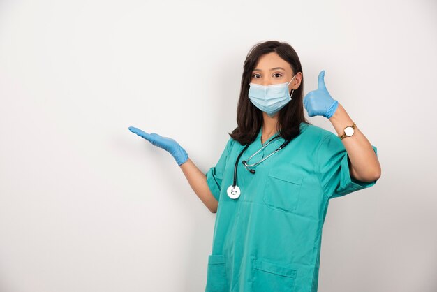 Молодой врач в медицинской маске и перчатках, делая большие пальцы руки вверх на белом фоне. Фото высокого качества