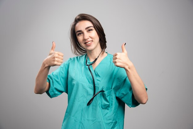 Young doctor in uniform giving thumbs up on gray wall.