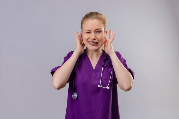 Young doctor wearing purple medical gown and stethoscope closed ears on isolated white wall