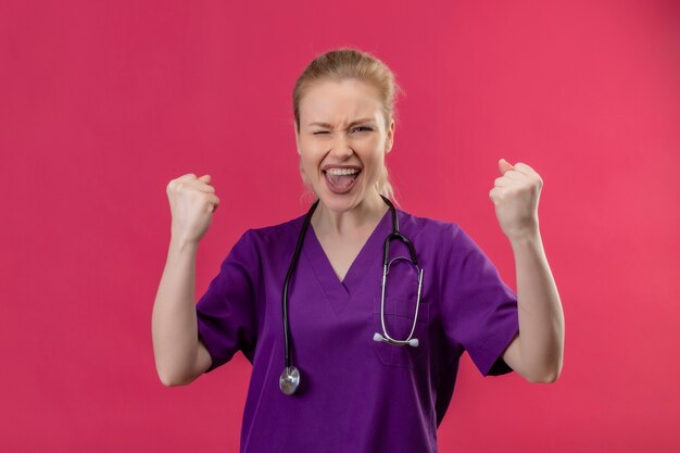 Young doctor wearing purple medical gown and stethoscope doing strong gesture on isolated pink wall