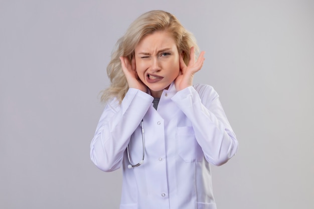 Young doctor wearing stethoscope in medical gown closed ears on white wall