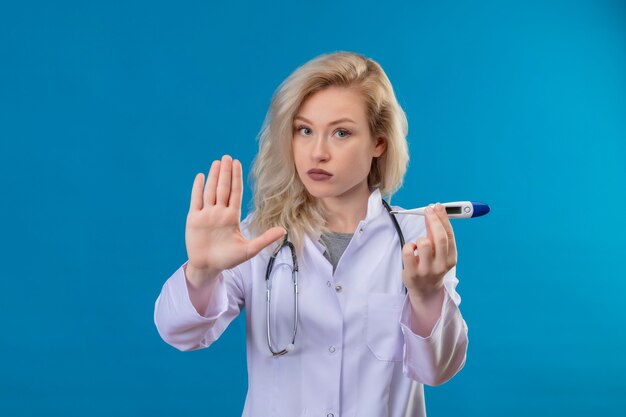 Young doctor wearing stethoscope in medical gown holding thermometer showing stop gesture on blue wall