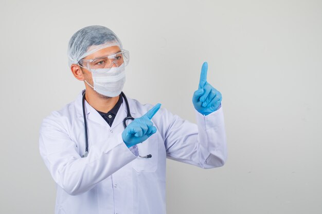 Молодой врач в белом халате, шляпе, перчатках указывает обеими пальцами рук