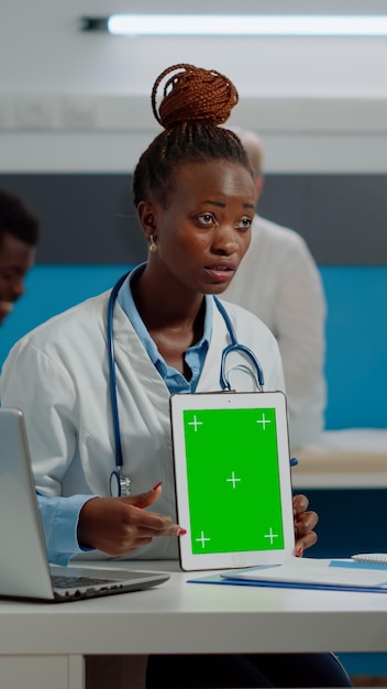 Young doctor with green screen technology on tablet explaining chroma key display with mockup template to elder patient. Medic vertically holding isolated background sitting at desk