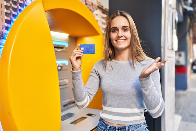 Молодая женщина-врач, держащая кредитную карту в банкомате, празднует достижение со счастливой улыбкой и выражением лица победителя с поднятой рукой
