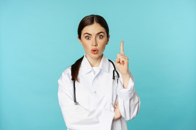 若い医者、白衣を着た女性医師が指を上げ、上向きに、smthを示唆し、解決策、啓示、トルコイズの背景の上に立っている