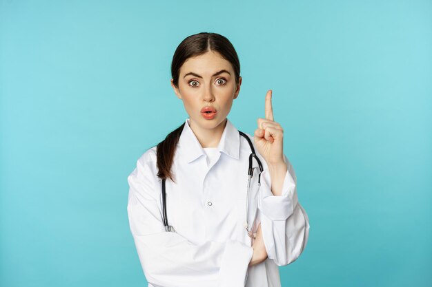 Young doctor, woman physician in white coat raising finger, pointing up, suggesting smth, has solution, revelation, standing over torquoise background