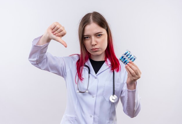 聴診器の医療ローブを身に着けている若い医者の女性は、孤立した白い壁に親指を下に丸薬を保持します