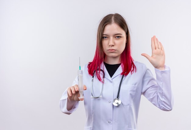 孤立した白い壁に停止ジェスチャーを示す注射器を保持している聴診器医療ローブを身に着けている若い医者の女性