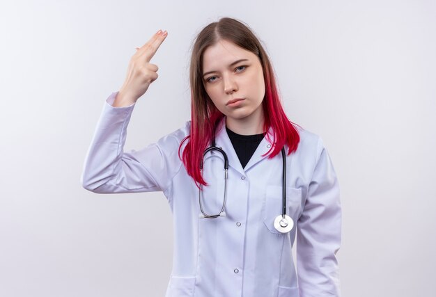 孤立した白い壁にピストルジェスチャーを示す聴診器医療ローブを身に着けている若い医者の女性