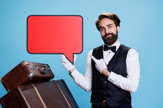 Free photo young doorman points at speech bubble against blue background, doing marketing ad using red billboard sign with copyspace. concierge employee holding blank isolated cardboard on camera.