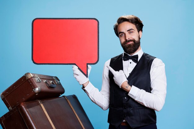 Young doorman points at speech bubble against blue background, doing marketing ad using red billboard sign with copyspace. Concierge employee holding blank isolated cardboard on camera.