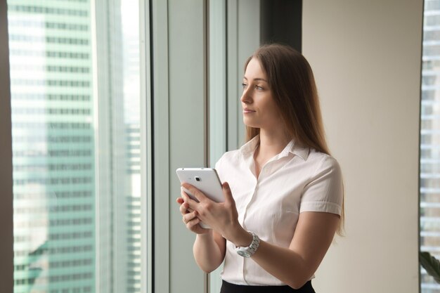 Young dreamy beautiful businesswoman looking through window while holding tablet