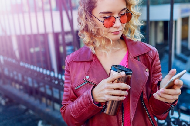 Foto gratuita giovane donna bionda vaga che cammina nella città e che per mezzo dello smart phone. dettagli da vicino. ragazza moderna alla moda con caffè. peli ventosi.