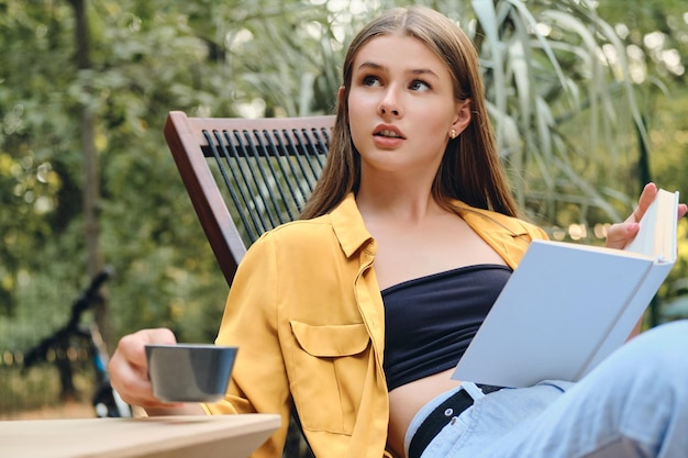 Young dreamy brown haired teenage girl in yellow shirt and top thoughtfully sitting with book and cup of coffee on wooden deck chair in city park
