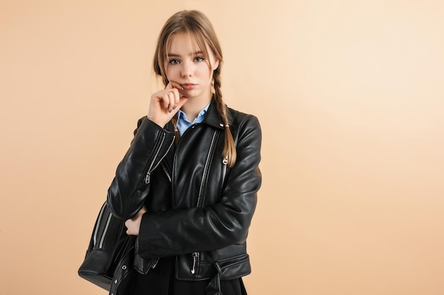 Young dreamy girl with two braids in leather jacket with black backpack on shoulder holding hand near face thoughtfully looking in camera over beige background isolated