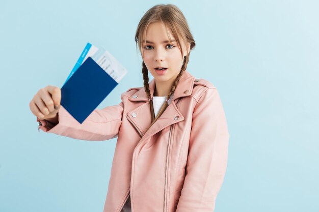 Young dreamy girl with two braids in pink leather jacket and white t-shirt showing passport with tickets thoughtfully looking in camera over blue background isolated