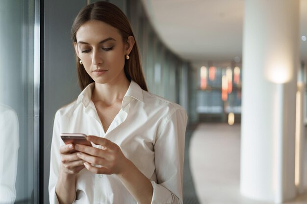 Young elegant lady in white collar shirt checking messages answering business partner during meeting break standing in office corridor using smartphone texting coworker rehearsing speech