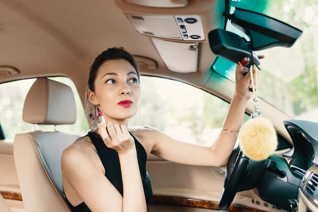 Young elegant woman looking in the car view mirror while applyin