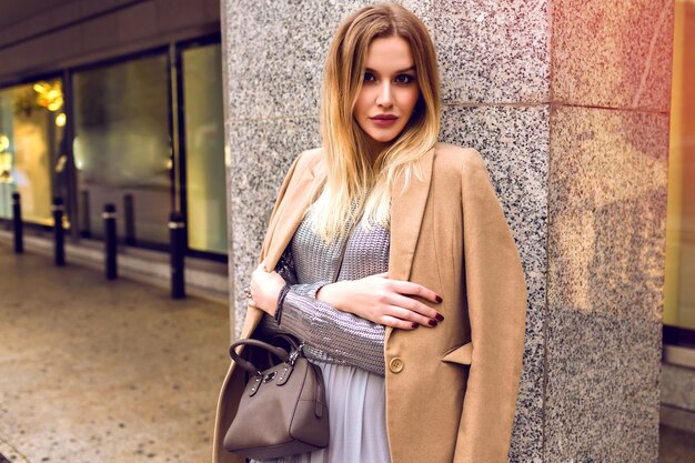 Young elegant woman posing on the street near shopping center, glamour trendy outfits, beige coat , silver sweater and dress, spring time, natural beauty