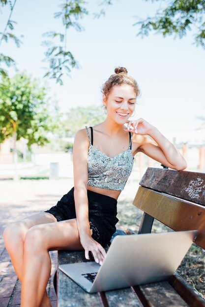 Young elegant woman with hat in white dress sitting on bench in park and working on laptop