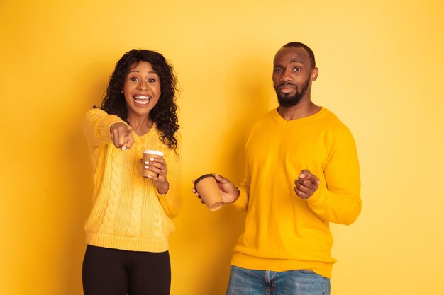 Young emotional african-american man and woman in bright casual clothes on yellow background. Beautiful couple. Concept of human emotions, facial expession, relations, ad. Drink coffee and pointing.