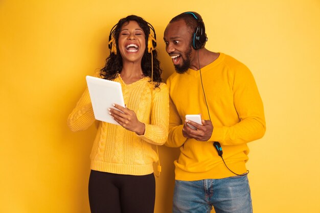 노란색 배경에 밝은 캐주얼 옷에 젊은 감정적 인 아프리카 계 미국인 남자와 여자. 아름다운 커플. 인간의 감정, 얼굴 expession, 관계, 광고의 개념. 태블릿 및 스마트 폰 사용.