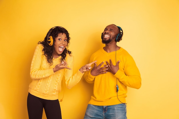 黄色の背景に明るいカジュアルな服を着た若い感情的なアフリカ系アメリカ人の男性と女性。美しいカップル。人間の感情、顔の表現、関係の概念。彼女は歌う男を指しています。